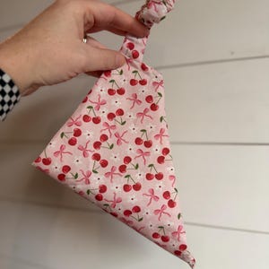 May include: A pink fabric item with a cherry and bow pattern. The fabric is a light pink with red cherries, pink bows, and small white flowers. The item is held up by a knotted fabric handle.