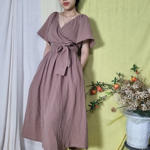 Puede incluir: Un vestido cruzado de color malva con mangas cortas y con volantes y un cinturón a juego. El vestido tiene un escote en V y una falda amplia. La modelo lleva una diadema blanca. El vestido está hecho de una tela texturizada.