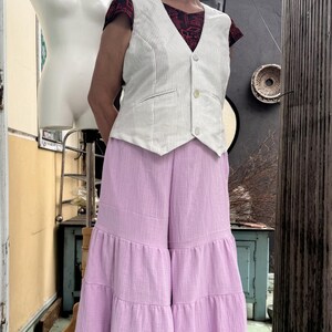 May include: A woman wearing a white pinstripe vest over a red patterned top, paired with wide-leg, tiered lavender pants. The outfit is a blend of textures and colors, creating a unique ensemble.