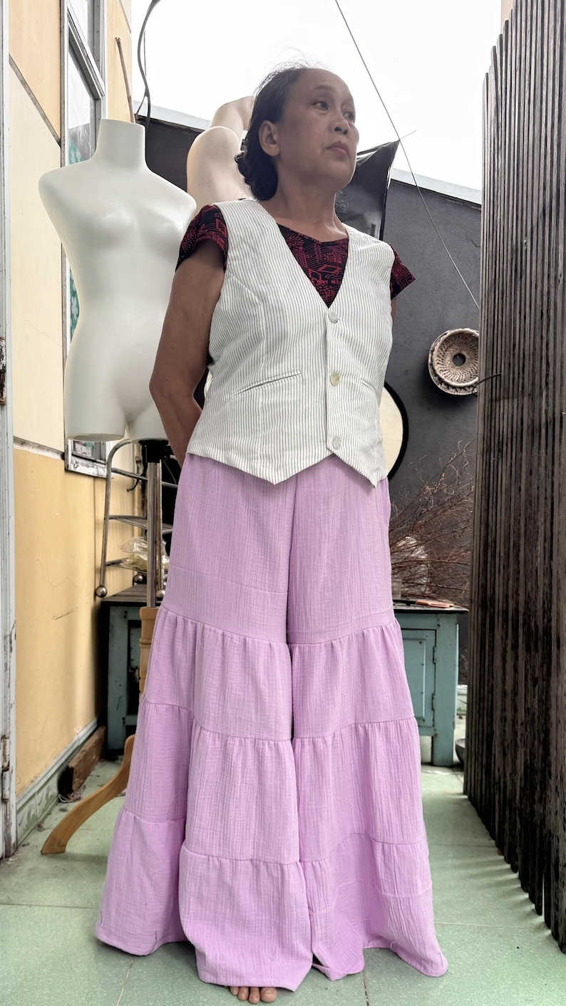 May include: A woman wearing a white pinstripe vest over a red patterned top, paired with wide-leg, tiered lavender pants. The outfit is a blend of textures and colors, creating a unique ensemble.