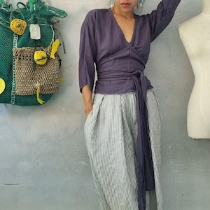 May include: A woman wearing a dark purple wrap top with a matching waist tie and striped pants. The top has three-quarter sleeves and a V-neck. The pants are white with vertical gray stripes. A green mesh bag with yellow accents hangs on the wall.