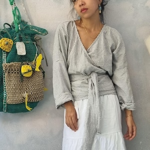 May include: A wrap-style top with long sleeves and a striped pattern in gray and white. The top is tied at the waist with a matching fabric belt. A white tiered skirt is also visible. A green mesh bag with yellow embellishments hangs on the wall.