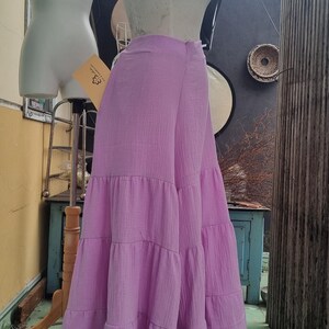May include: A light purple, tiered maxi skirt displayed on a mannequin. The skirt has a flowing, layered design and is made of a textured fabric. The mannequin is positioned in front of a neutral background.