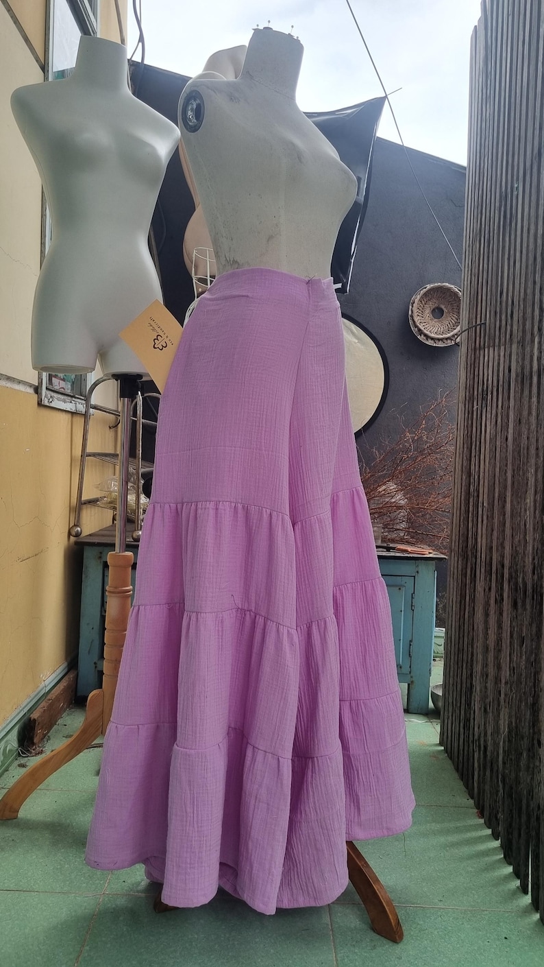 May include: A light purple, tiered maxi skirt displayed on a mannequin. The skirt has a flowing, layered design and is made of a textured fabric. The mannequin is positioned in front of a neutral background.