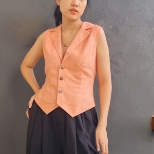 May include: A sleeveless peach-colored vest with a notched collar and three brown buttons. The vest is paired with black wide-leg pants. The outfit is simple and elegant.