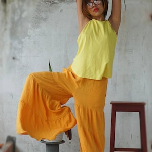 May include: A yellow halter top and orange palazzo pants. The outfit is made of a textured fabric. The model is wearing glasses and has their arm raised. A small stool and a plant are in the background.