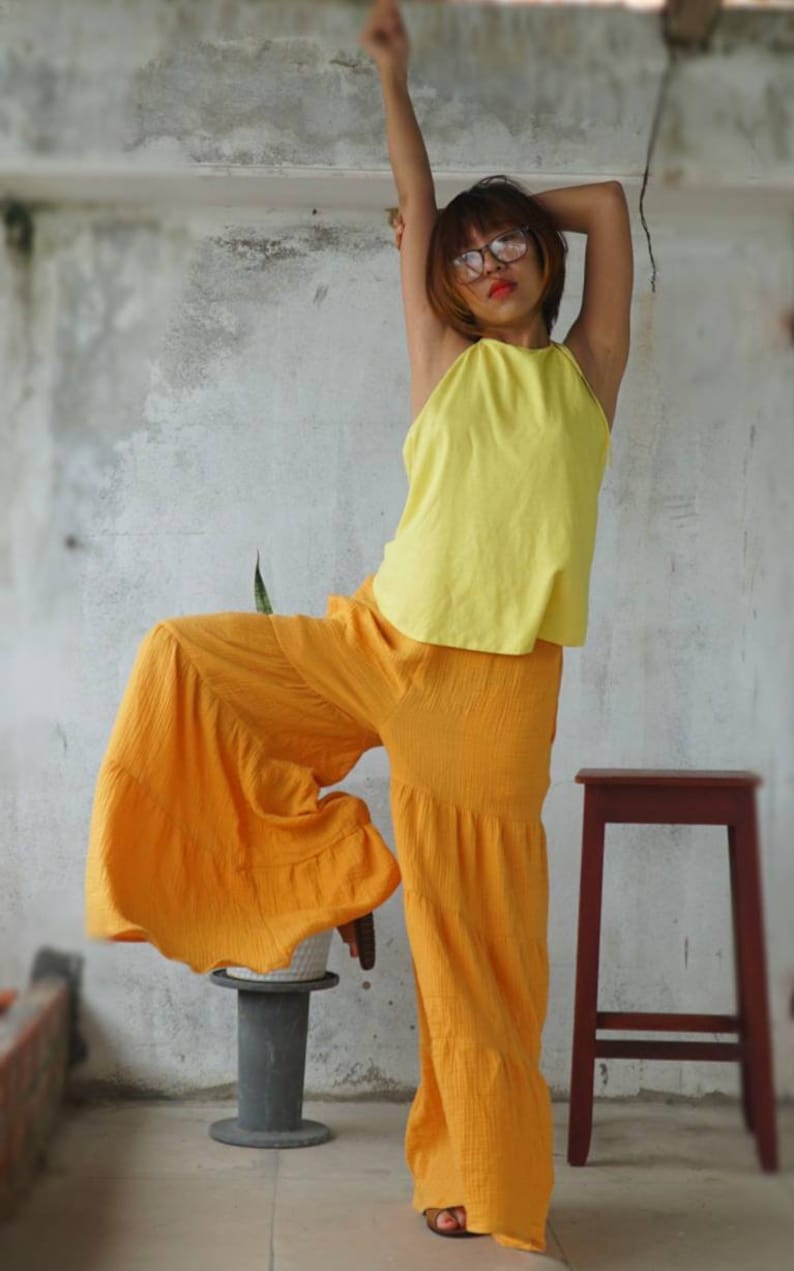 May include: A yellow halter top and orange palazzo pants. The outfit is made of a textured fabric. The model is wearing glasses and has their arm raised. A small stool and a plant are in the background.