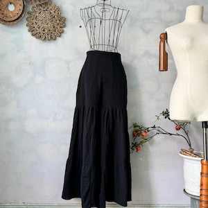 May include: A long, black tiered skirt displayed on a wire dress form. The skirt has a high waist and a flowing, layered design. The fabric appears to be a lightweight material, suitable for warmer weather. The skirt is styled on a mannequin.
