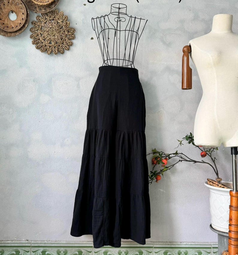 May include: A long, black tiered skirt displayed on a wire dress form. The skirt has a high waist and a flowing, layered design. The fabric appears to be a lightweight material, suitable for warmer weather. The skirt is styled on a mannequin.