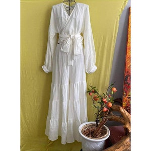 May include: A white, long-sleeved wrap top and tiered pants set. The outfit is tied at the waist with a matching fabric belt. A small potted plant with orange fruit is in the foreground.