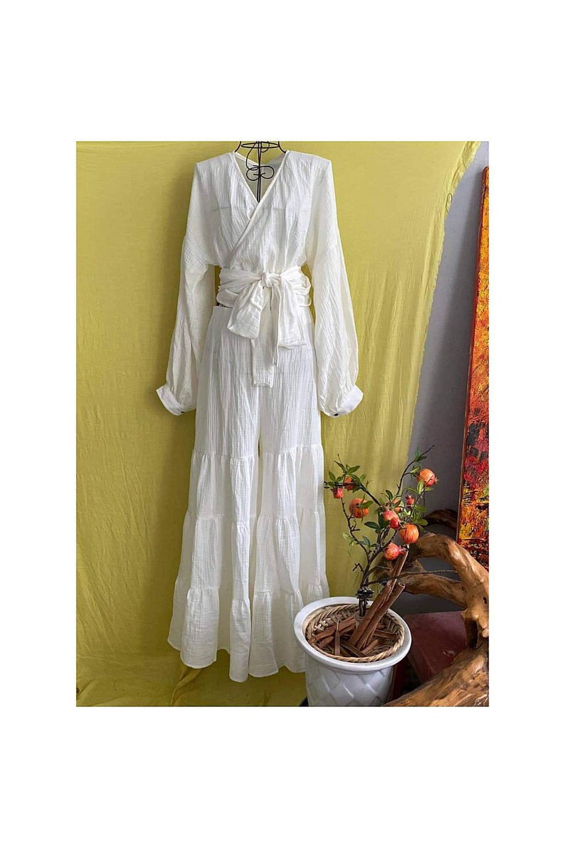 May include: A white, long-sleeved wrap top and tiered pants set. The outfit is tied at the waist with a matching fabric belt. A small potted plant with orange fruit is in the foreground.