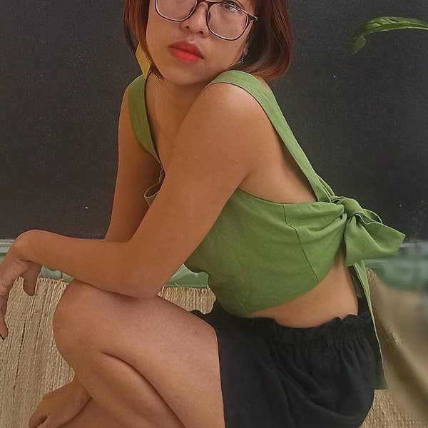 May include: A sleeveless olive green crop top featuring wide shoulder straps and a large fabric tie closure at the back, paired with black shorts that have an elasticized waistband. The top is constructed with a simple, solid-colored design.