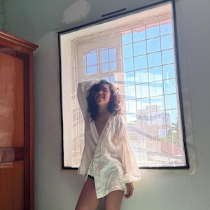 May include: A white long-sleeved wrap blouse with a deep V-neckline and voluminous sleeves, featuring a lightweight, semi-sheer fabric. The garment is worn over dark bottoms, showcasing a relaxed fit with a slightly flared hemline and cuffs.