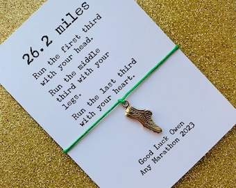 May include: A white card with the text "26.2 miles" and a poem about running a marathon. A small gold-colored shoe charm hangs from a green string. The card is on a gold glitter surface.