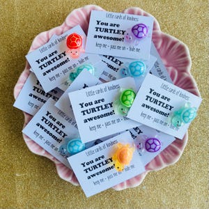 May include: A collection of small white rectangular cards featuring the text Little cards of kindness, You are TURTLEY awesome!, and keep me - pass me on - hide me. Each card is adorned with a small, colourful, translucent plastic turtle figurine.