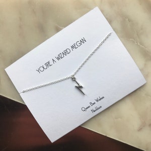 May include: A silver lightning bolt charm necklace on a white card. The card has the text "YOU'RE A WIZARD MEGAN" and "Queen Bee Wishes Necklace". The background is a marble surface.
