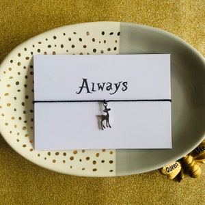 May include: A white card with the word "Always" printed in black script, with a black string bracelet and a silver deer charm. The card is on a decorative plate with gold polka dots and a gold background.
