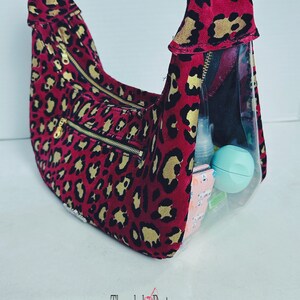 Puede incluir: Un bolso de mano de leopardo rojo y dorado con una bolsa transparente con cremallera en la parte delantera. La bolsa está llena de varios artículos, incluyendo una pequeña botella, un recipiente redondo y una pequeña libreta.