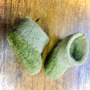 以下が含まれることがあります： ふわふわの質感の緑色のフェルトスリッパ2足。スリッパは木製の表面の上に置かれています