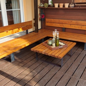 Pu&ograve; includere: Un set di sedute da esterno in legno con una panca ad angolo e un tavolino quadrato. La panca ha una finitura in legno naturale e il tavolino ha una tinta marrone scuro. Il set &egrave; su un ponte in legno.