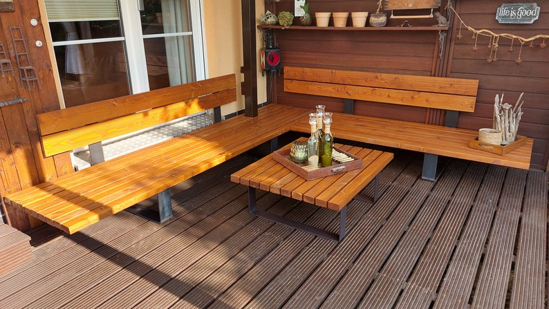 Pu&ograve; includere: Un set di sedute da esterno in legno con una panca ad angolo e un tavolino quadrato. La panca ha una finitura in legno naturale e il tavolino ha una tinta marrone scuro. Il set &egrave; su un ponte in legno.
