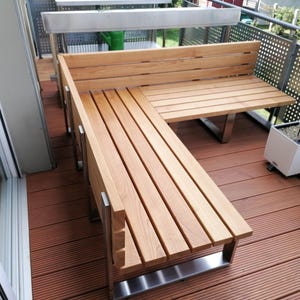 Könnte beinhalten: Eine Eckbank aus Holz mit einem Gestell aus Edelstahl. Die Bank steht auf einem Holzdeck mit einer Topfpflanze im Hintergrund.