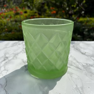 May include: A light green glass tumbler with a diamond pattern. The glass has a thick base and a slightly flared rim. The tumbler is on a white and grey marble surface.