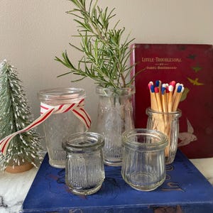 May include: Several small, clear glass jars in different shapes and sizes. One jar is tied with a red and white striped ribbon. Some jars contain rosemary sprigs and matchsticks. A red book with gold lettering is in the background.