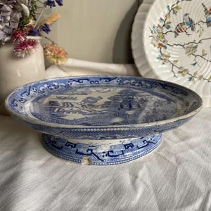 May include: A vintage blue and white ceramic cake stand. The stand features a detailed landscape design with a pagoda and other architectural elements. The stand has a round base and a wide, flat top, ideal for displaying cakes or other items. The stand is approximately 25 cm in diameter.