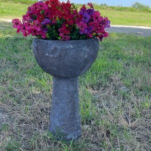 May include: A dark gray, stone-like planter filled with vibrant pink, red, and purple flowers. The planter sits on a pedestal in a grassy outdoor setting. The flowers are in full bloom, adding a splash of color.
