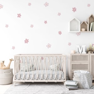 May include: A white crib with a wicker frame and a white mattress. The crib is in a nursery with a white wall decorated with pink flower decals. There is a wooden dresser with a white blanket basket and a wooden shelf with a white house-shaped box on top.