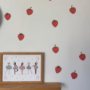 May include: A framed print of five ballerinas in pink and grey tutus with a white background and pink stars. The print is on a wooden shelf with three hooks. The wall behind the shelf is decorated with red strawberry stickers.