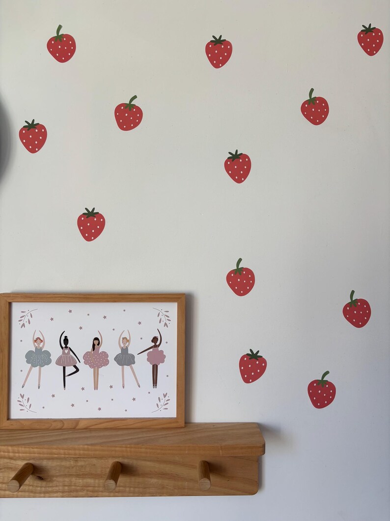 May include: A framed print of five ballerinas in pink and grey tutus with a white background and pink stars. The print is on a wooden shelf with three hooks. The wall behind the shelf is decorated with red strawberry stickers.