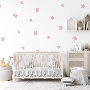 May include: A white crib with a white and gray patterned mattress and a white wicker basket with a stuffed animal and a giraffe toy. The crib is in front of a white wall with pink flower decals. There is a wooden dresser with a white basket and a wooden shelf with a white house shaped decor.