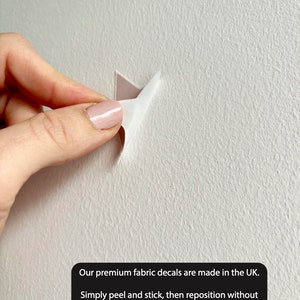 May include: A hand peeling back a corner of a fabric decal to show the adhesive backing. The decal is being applied to a white wall. The text reads: "Our premium fabric decals are made in the UK. Simply peel and stick, then reposition without leaving any marks or damage!"
