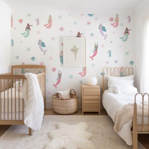 May include: A nursery with a mermaid theme. The white walls are decorated with pink, teal, and blue mermaid decals, starfish, and dots. The room features a wooden crib, a bed, a woven basket, a framed picture, and a white rug.