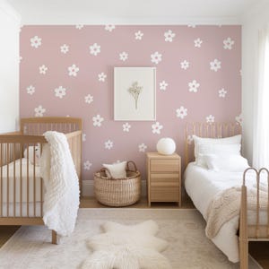 May include: Two cribs with white bedding in a nursery with a pink wall featuring a white daisy pattern. A white rug with a fluffy white sheepskin rug on top is in the center of the room.