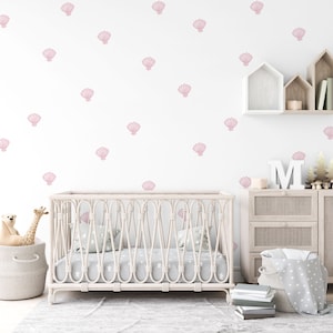 Può includere: Un lettino bianco con un materasso a motivi rosa e bianchi e un cesto di vimini bianco contenente un peluche e un giocattolo giraffa. La parete dietro il lettino è bianca con un motivo a conchiglie rosa.