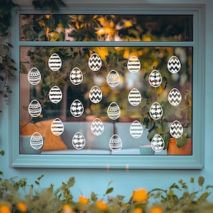 May include: A window with white Easter egg stickers on the glass. The stickers are in various patterns and designs. The window is framed in light blue paint.
