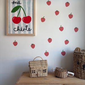 May include: A framed print with a blue and white striped background and a drawing of two red cherries with green stems and leaves. The text "ma ch&eacute;rie" is written below the cherries. The print is surrounded by red strawberry stickers on a white wall.