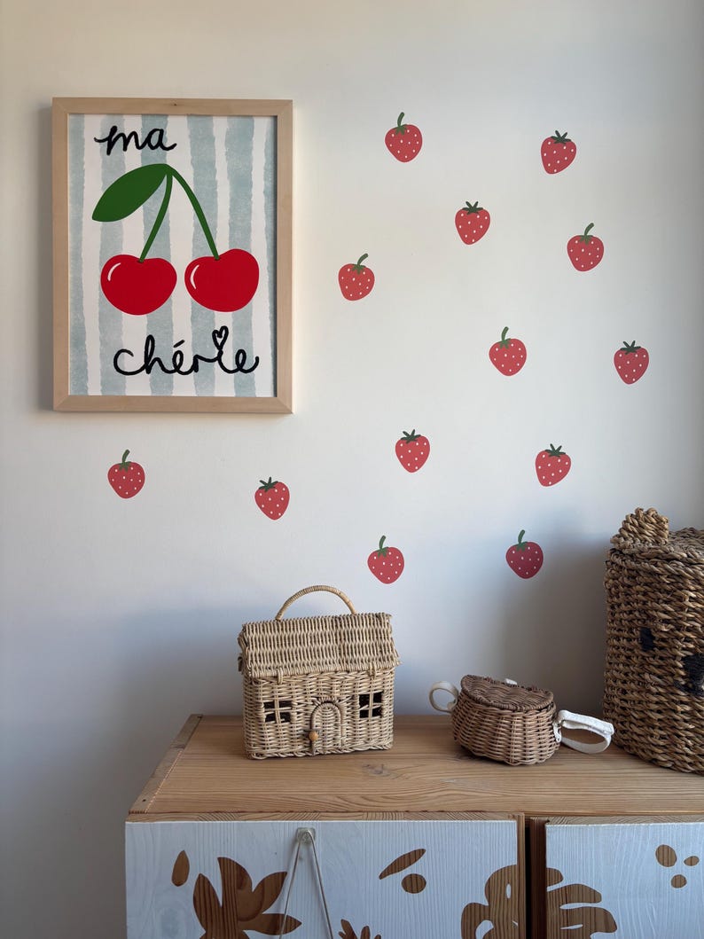 May include: A framed print with a blue and white striped background and a drawing of two red cherries with green stems and leaves. The text "ma ch&eacute;rie" is written below the cherries. The print is surrounded by red strawberry stickers on a white wall.
