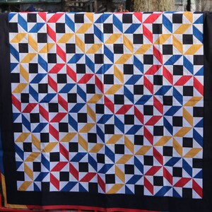 Puede incluir: Una colcha con un patrón geométrico de cuadrados y triángulos negros, blancos, rojos, azules y amarillos. La colcha está colgada de una valla metálica roja.