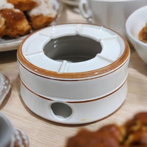 Könnte beinhalten: Weiße Keramik-Fondue-Schale mit braunem Rand und einem Loch im Boden. Die Schale steht auf einer Holzoberfläche.