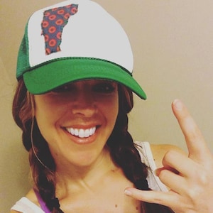 May include: A woman with long brown hair in braids is wearing a white and green trucker hat with a red and blue donut pattern. The hat has a green mesh back and a white front with a state outline.
