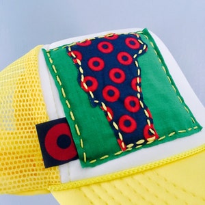 May include: Yellow trucker hat with a green, white, and blue patch featuring a red and blue circle pattern. The patch is sewn on with yellow stitching.