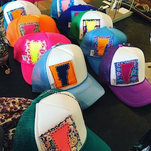Peut inclure: Une collection de casquettes de camionneur colorées avec une variété de patchs brodés représentant le contour de l'État du Vermont. Les casquettes sont en maille blanche avec une variété de bords et de patchs de couleurs.