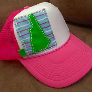 May include: A pink and white trucker hat with a green state outline sewn onto a blue and white striped patch. The hat has a mesh back.