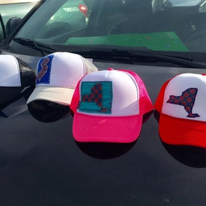 May include: Four trucker hats with a New York state outline embroidered on the front. The hats are black, white, pink, and red. The New York state outlines are all different colors and patterns.