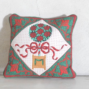 Peut inclure: Un coussin décoratif matelassé carré avec un panneau central en losange blanc orné d'un motif de topiaire de poinsettias rouges, d'un nœud en ruban rouge et d'un pot beige. La bordure est verte avec des motifs floraux rouges et un passepoil rouge.