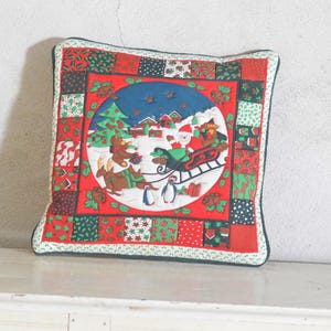 Puede incluir: Un cojín decorativo cuadrado con un borde de patchwork de tela con motivos navideños en rojo, verde y blanco. El panel central muestra una escena invernal con Santa en un trineo, un árbol de Navidad, un oso y pingüinos sobre la nieve.